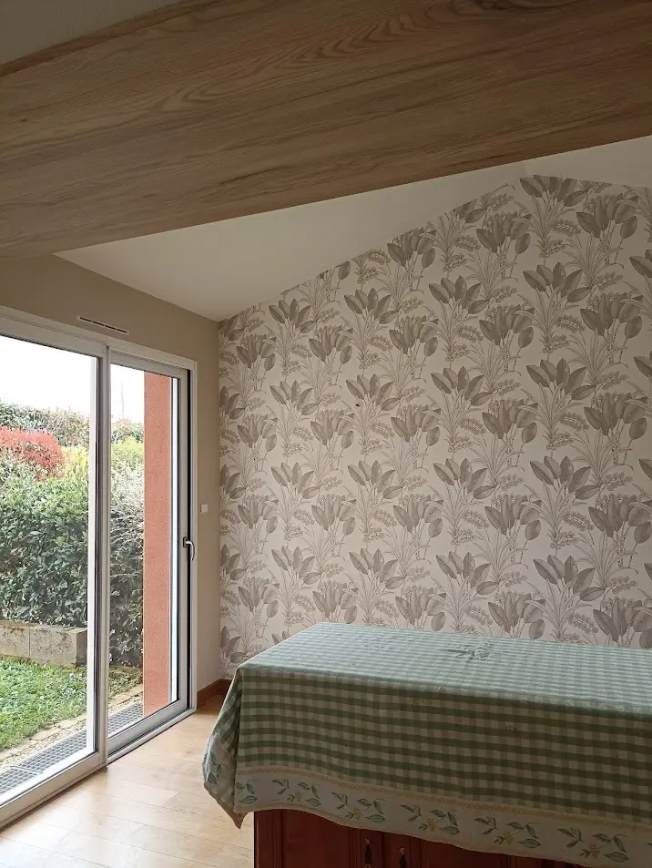 Travail de finition pour la pose de papier peint à motifs végétaux réalisé par Ramboz Le Peintre sur un mur sous rampant. L'image montre le raccord précis près de la baie vitrée et de la poutre en bois.
