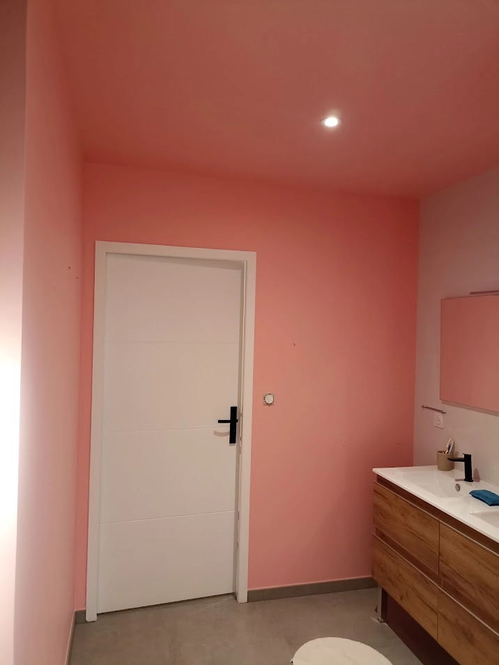 Vue des finitions de peinture rose corail dans une salle de bain réalisée par Ramboz Le Peintre. L'image montre le raccord net avec la porte blanche contemporaine et le meuble vasque en bois avec mitigeur noir.