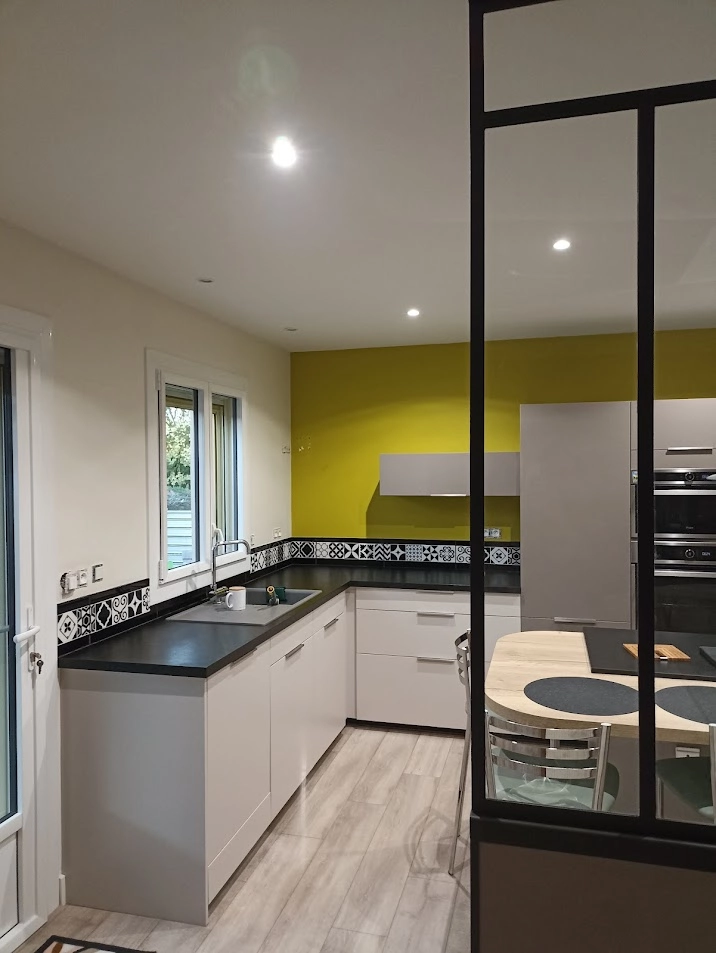Rénovation de peinture dans une cuisine moderne réalisée par Ramboz Le Peintre. Un mur d'accent de couleur vert sauge contraste avec les murs blancs, le plan de travail noir et la crédence en carreaux de ciment. Vue à travers une verrière d'atelier.
